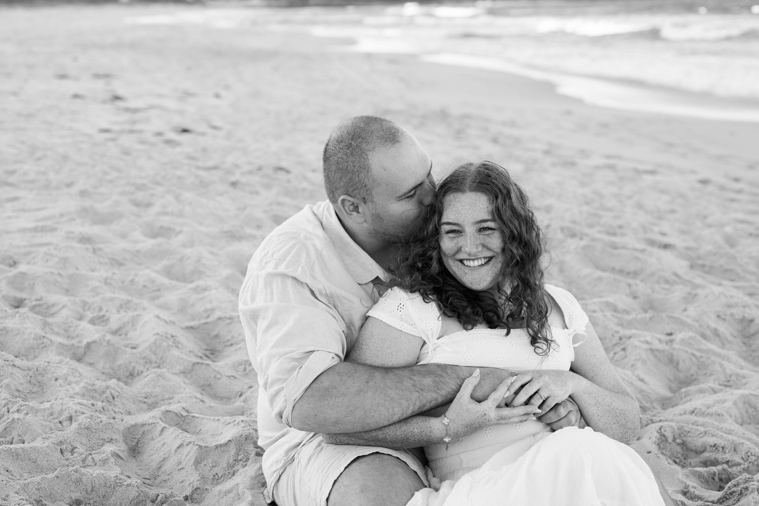 Couple sitting on beach sand in black and white photo, man kissing woman's cheek as she smiles warmly.