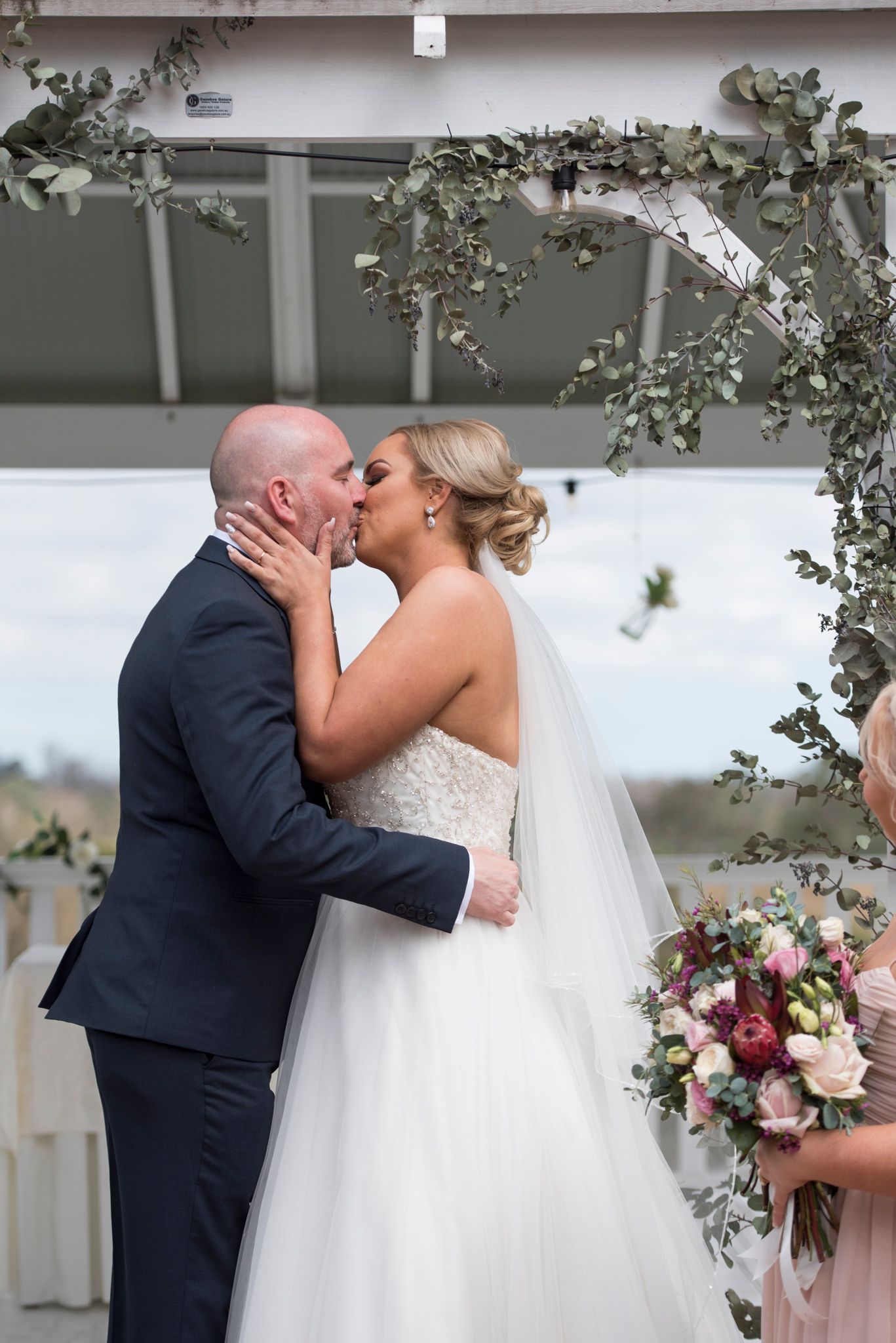 A romantic wedding couple shares a tender kiss under a white gazebo decorated with floral arrangements.