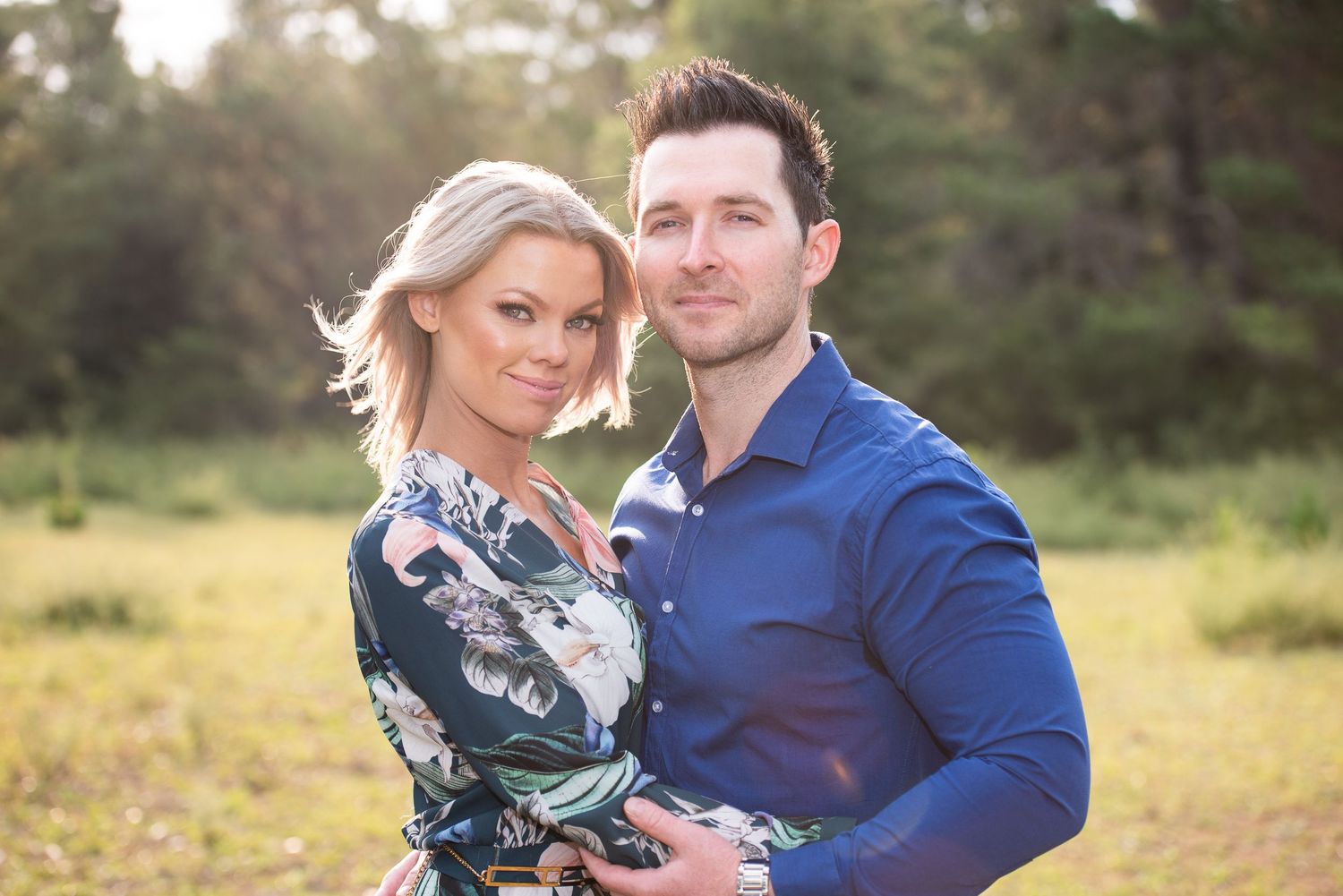 A couple poses together in a sunlit forest clearing.