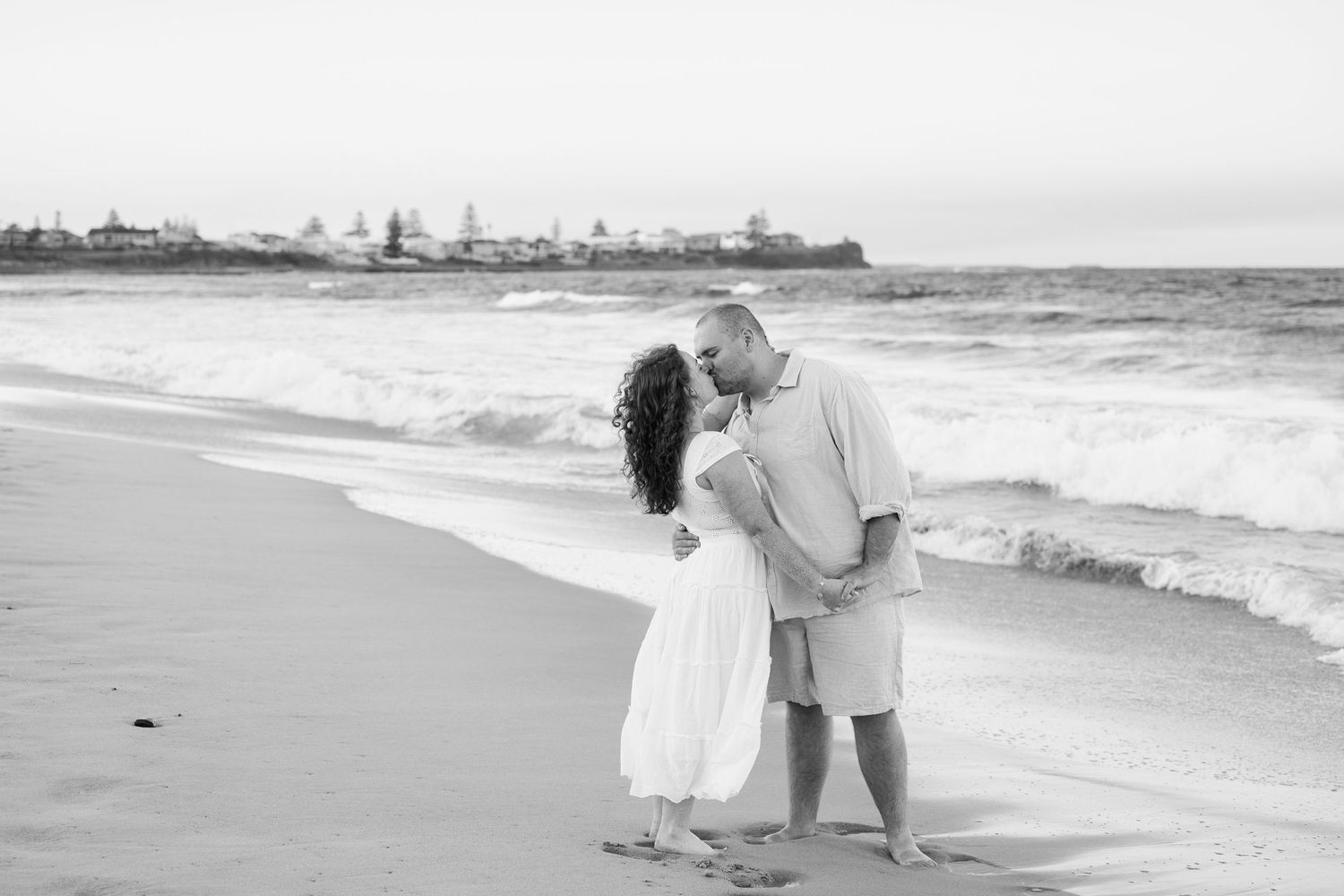 Black and white photo of a couple kissing on a sandy beach with ocean waves in the background.