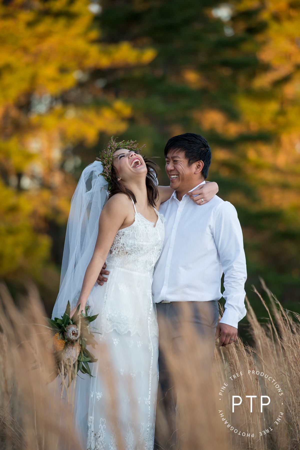 Wedding couple in white attire laughing together among tall autumn grasses at sunset.