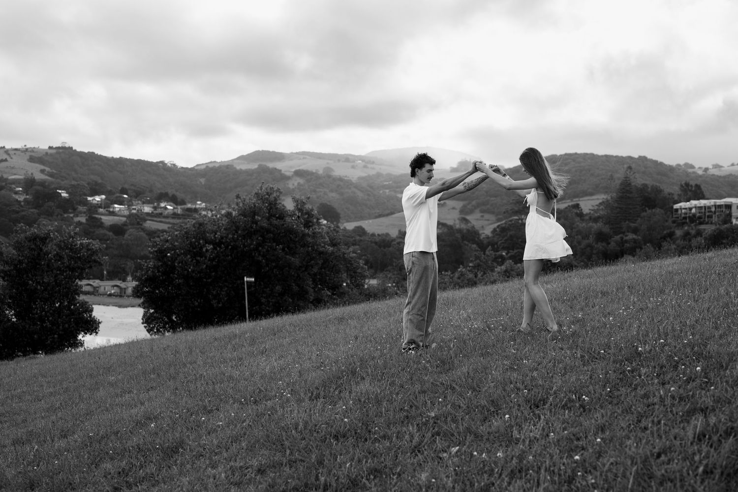 Two people stand in silhouette against rolling hills and mountains on a grassy slope at dusk.