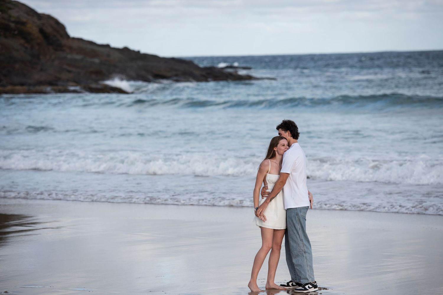 Couple sharing romantic moments along a scenic beach in Kiama with crashing waves and rocky coastline in the background.