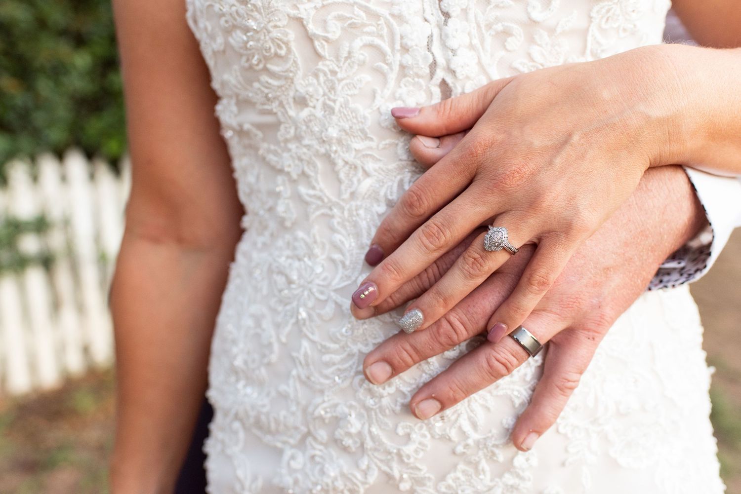 Detailed view of delicate white lace wedding dress with hands displaying rings.