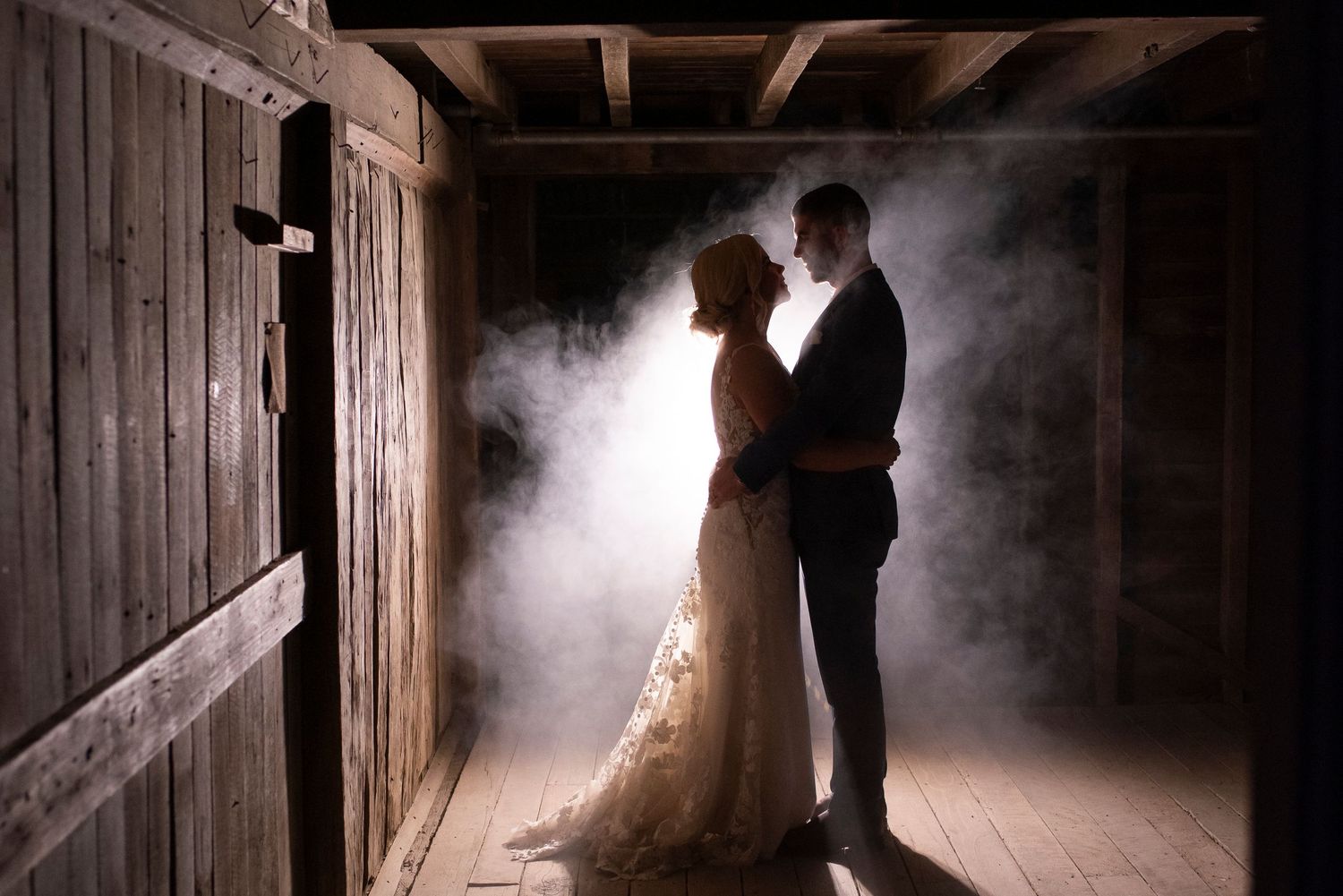 Silhouetted couple shares an intimate moment in a dimly lit barn with dramatic lighting and fog effects.