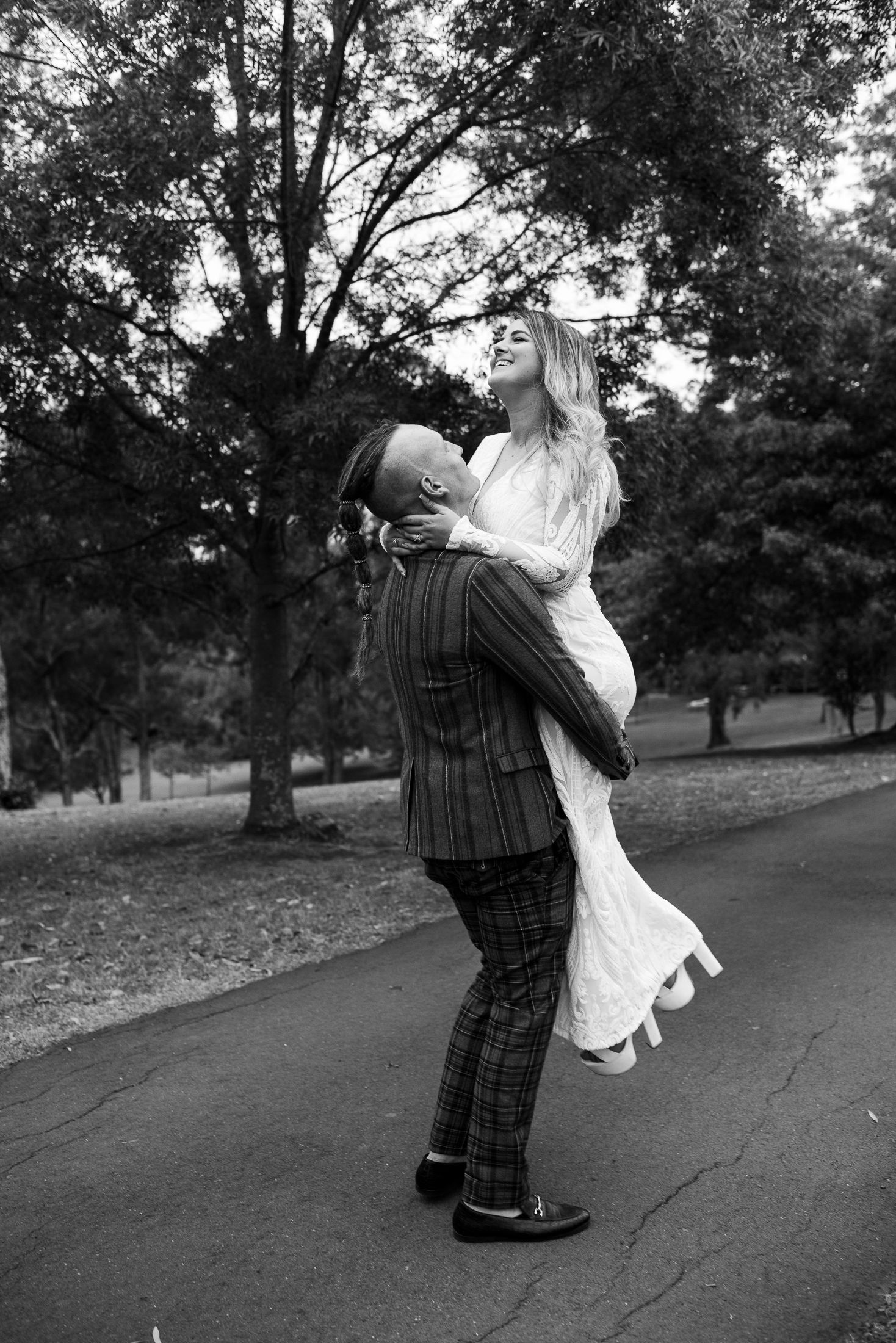 A romantic black and white photo of a bride and groom embracing in a park with trees in the background.