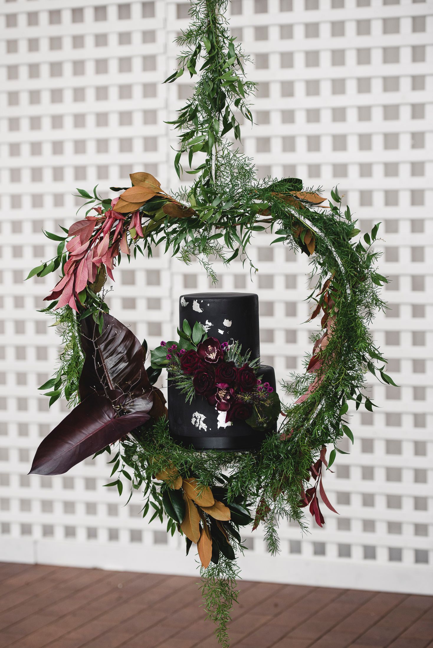 Modern black wedding cake display with cascading greenery and burgundy floral accents.