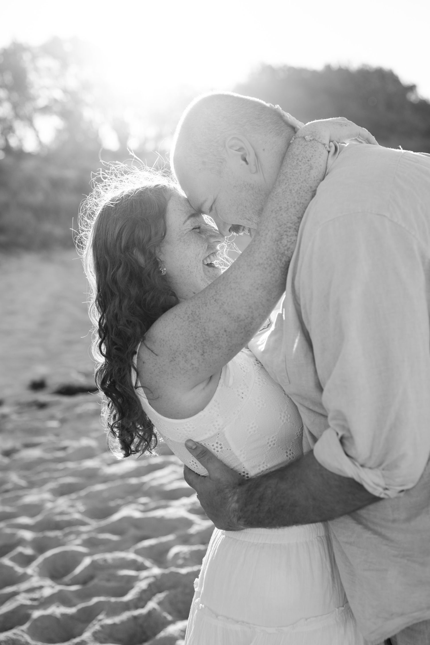Black and white photo of a couple sharing a romantic kiss on a sandy beach with sunlight glowing behind them.