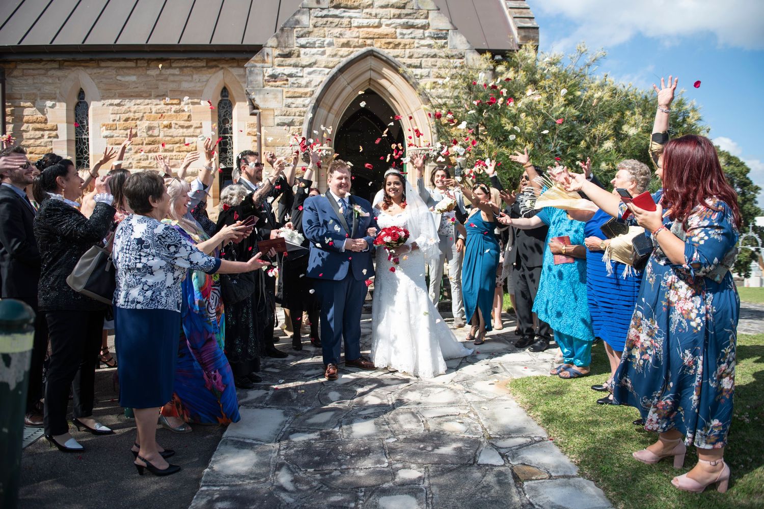 Wedding guests throw confetti and celebrate outside a historic stone church building during a joyous ceremony.