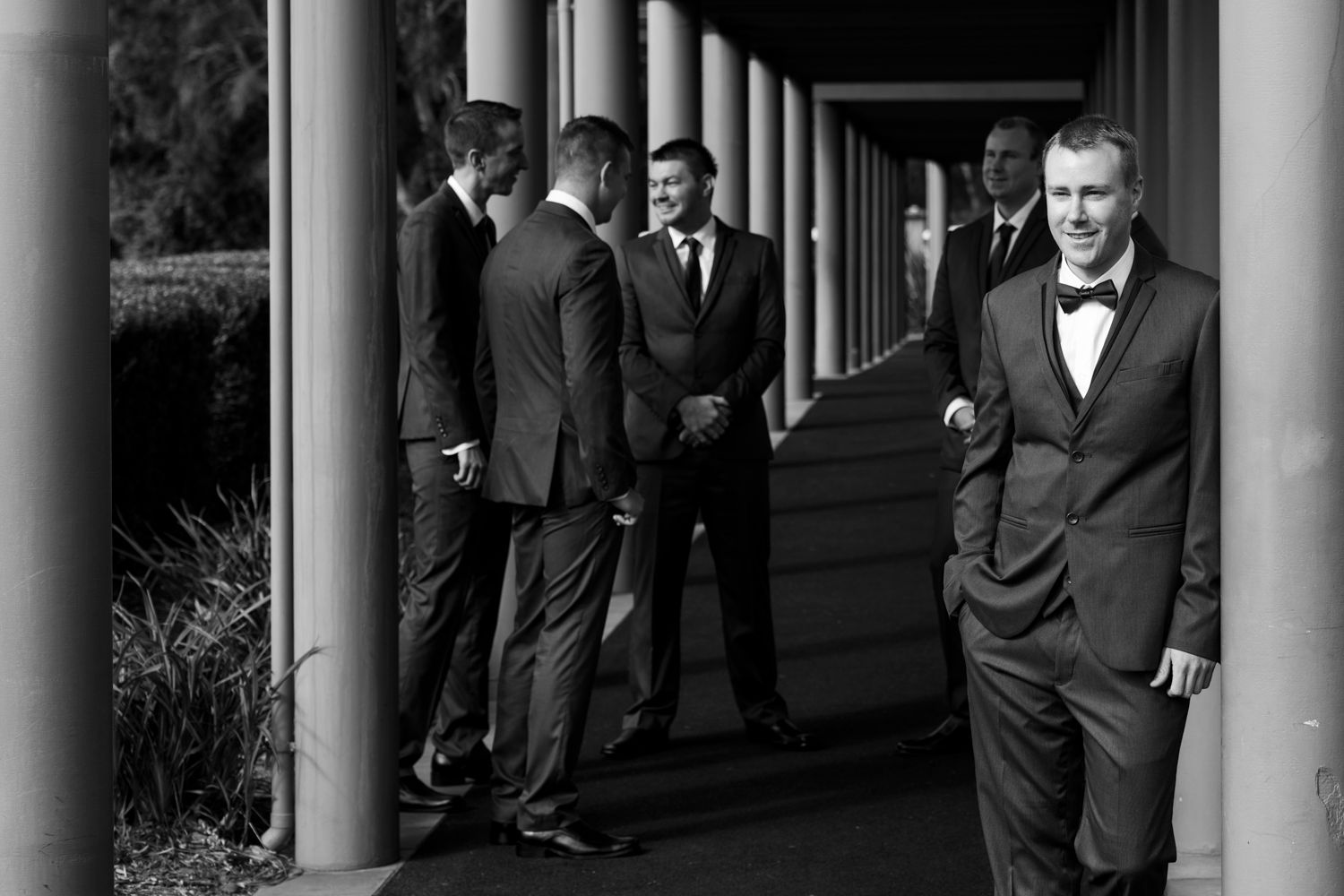 Groomsmen in black suits pose dramatically along a colonnade with white pillars.