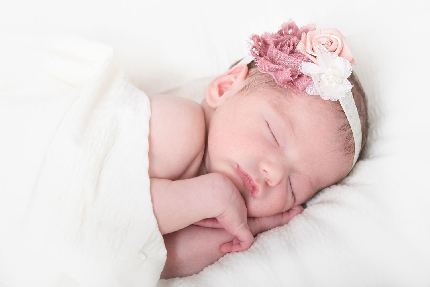 Peaceful sleeping newborn with pink headband resting on white blanket.