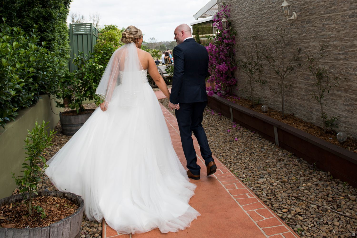 A couple walks along a brick path surrounded by lush greenery and stone walls at a wedding venue.