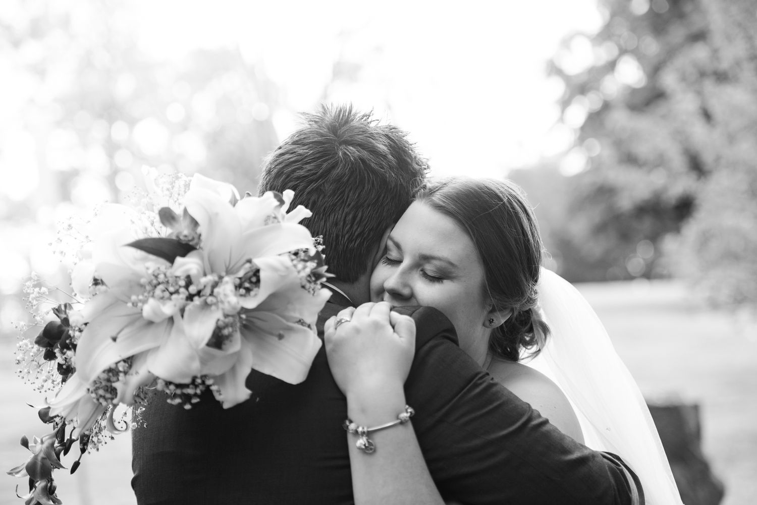 A tender black and white wedding moment captured outdoors as a couple embraces with a beautiful floral bouquet.