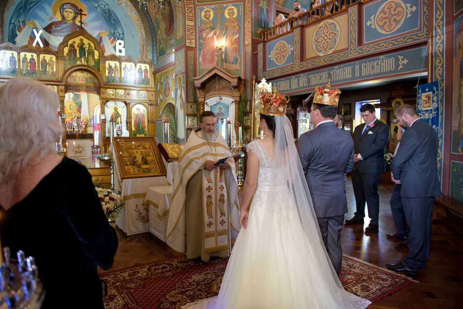 Wedding ceremony taking place inside a Russian Orthodox church with gilded icons and decorated walls and ceiling.