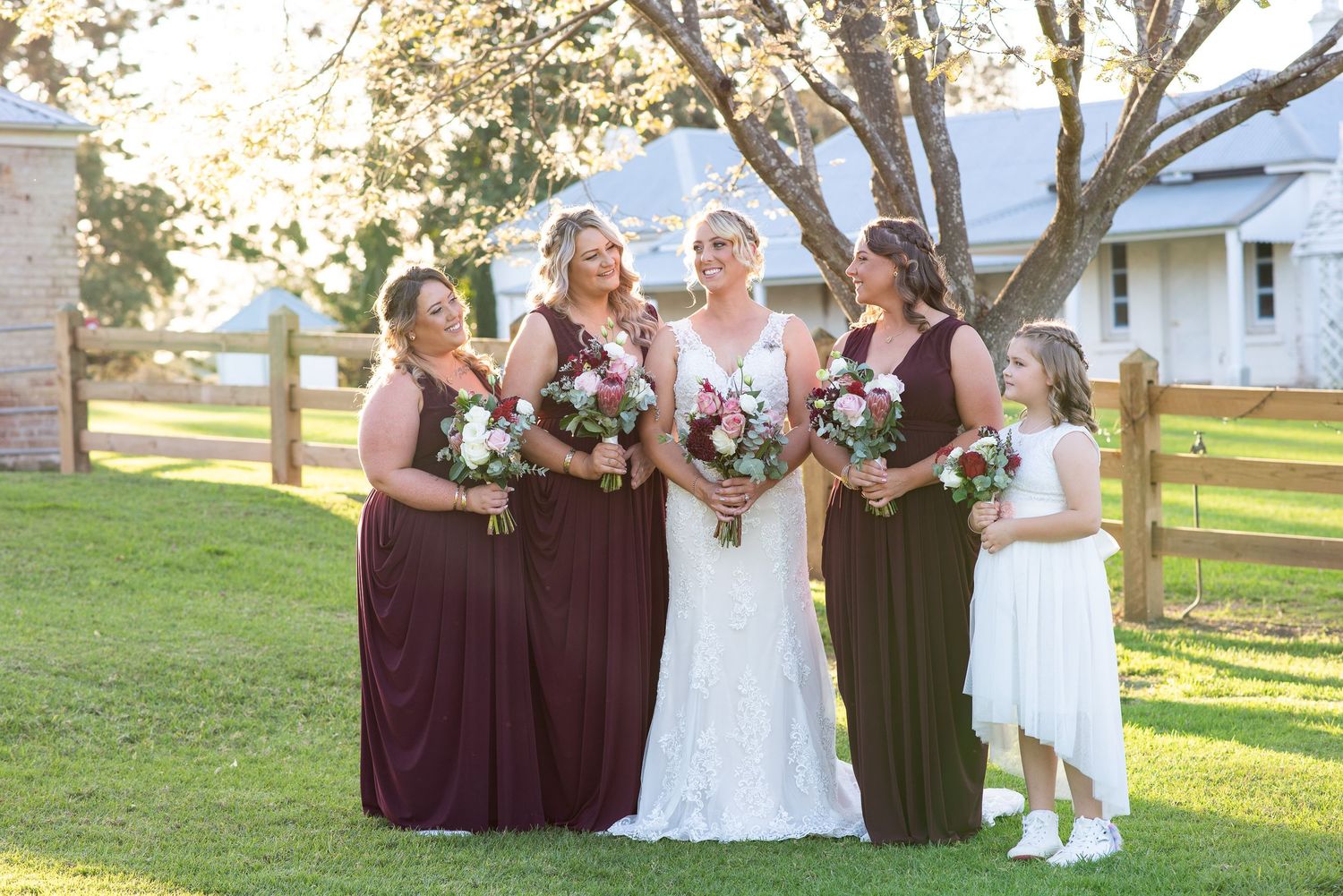 Brid with bridesmaids in burgundy dresses poses together on a sunny lawn with bouquets near a white building.