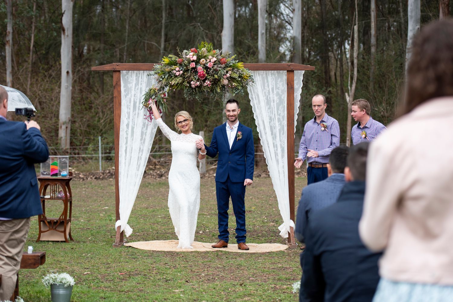 Newlyweds celebrate under a white draped arch during an outdoor bush wedding ceremony.