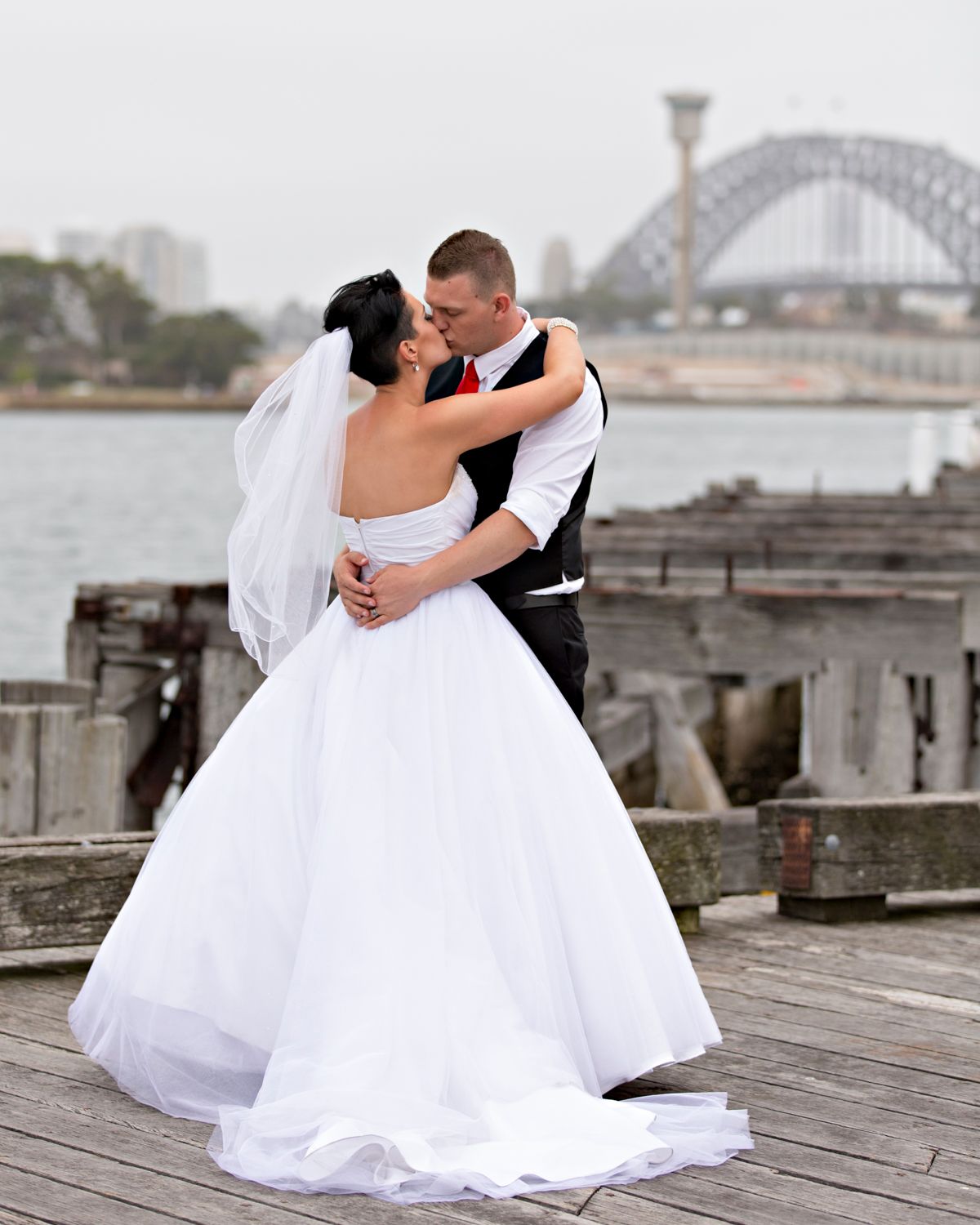 Romantic wedding portraits on wooden pier with harbour bridge visible in background.