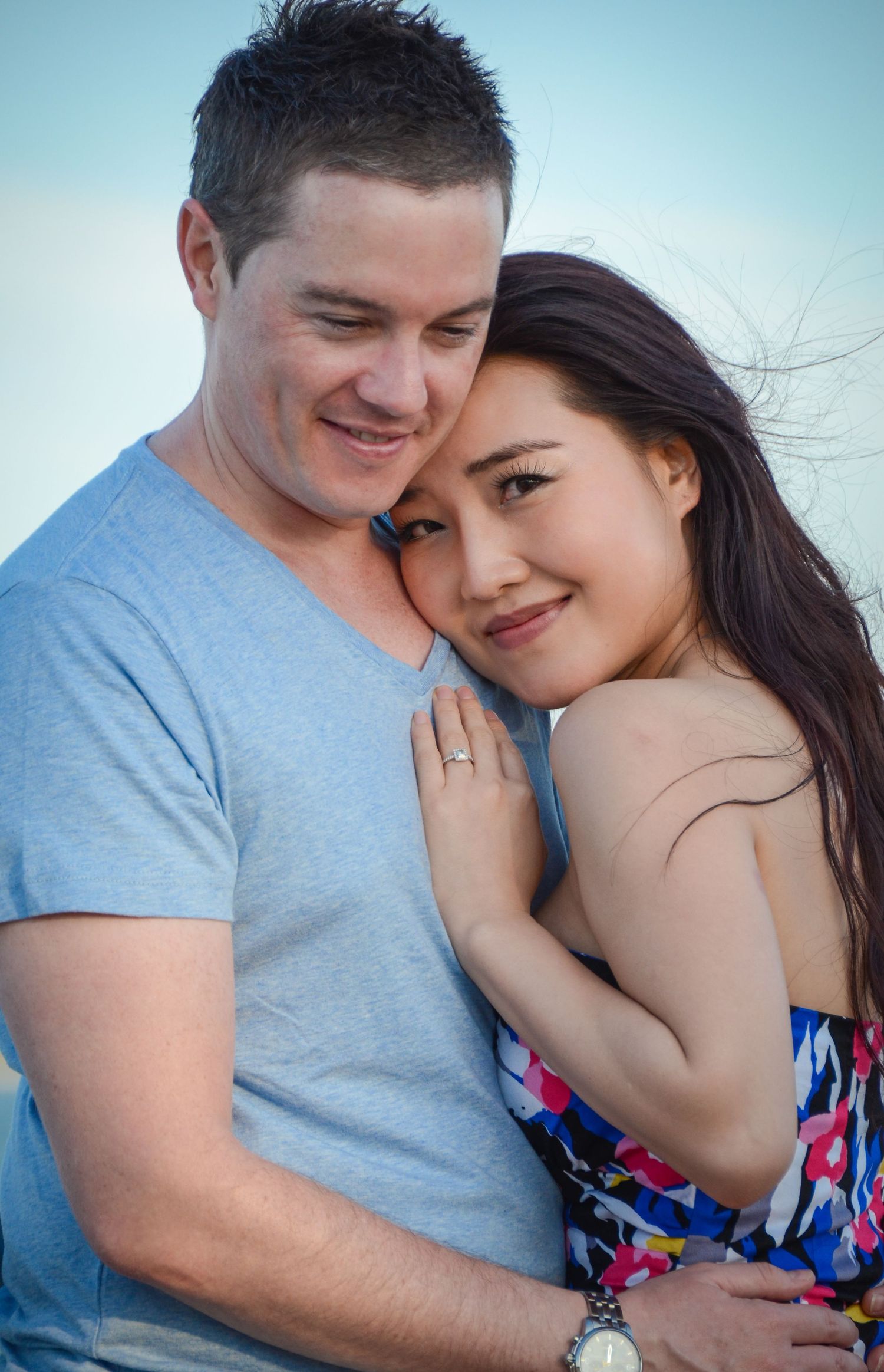 Tender embrace between couple wearing blue shirt and floral dress against bright sky.