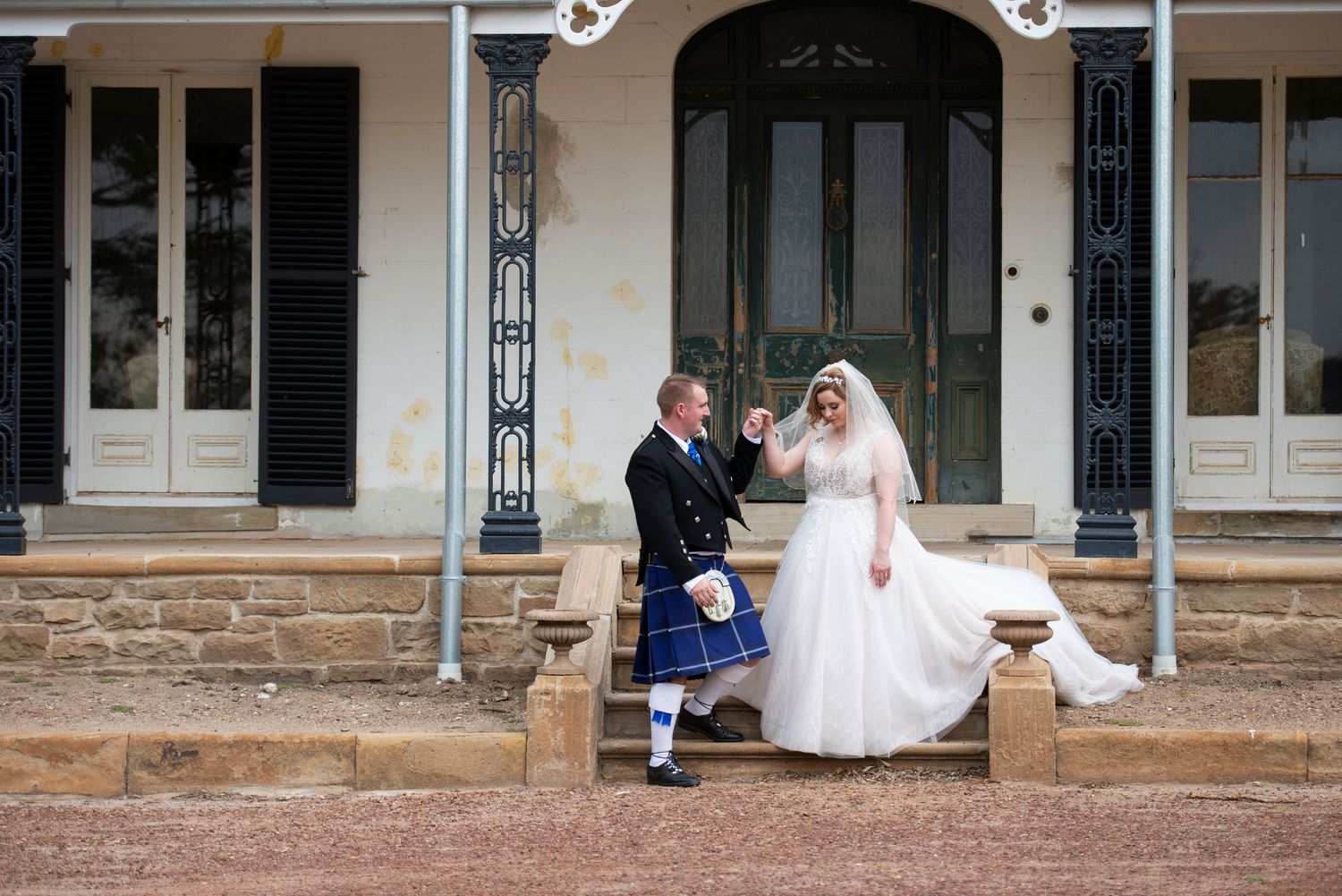 Bride and groom in Scottish kilt celebrate outside historic colonial building, sharing a joyful wedding moment.