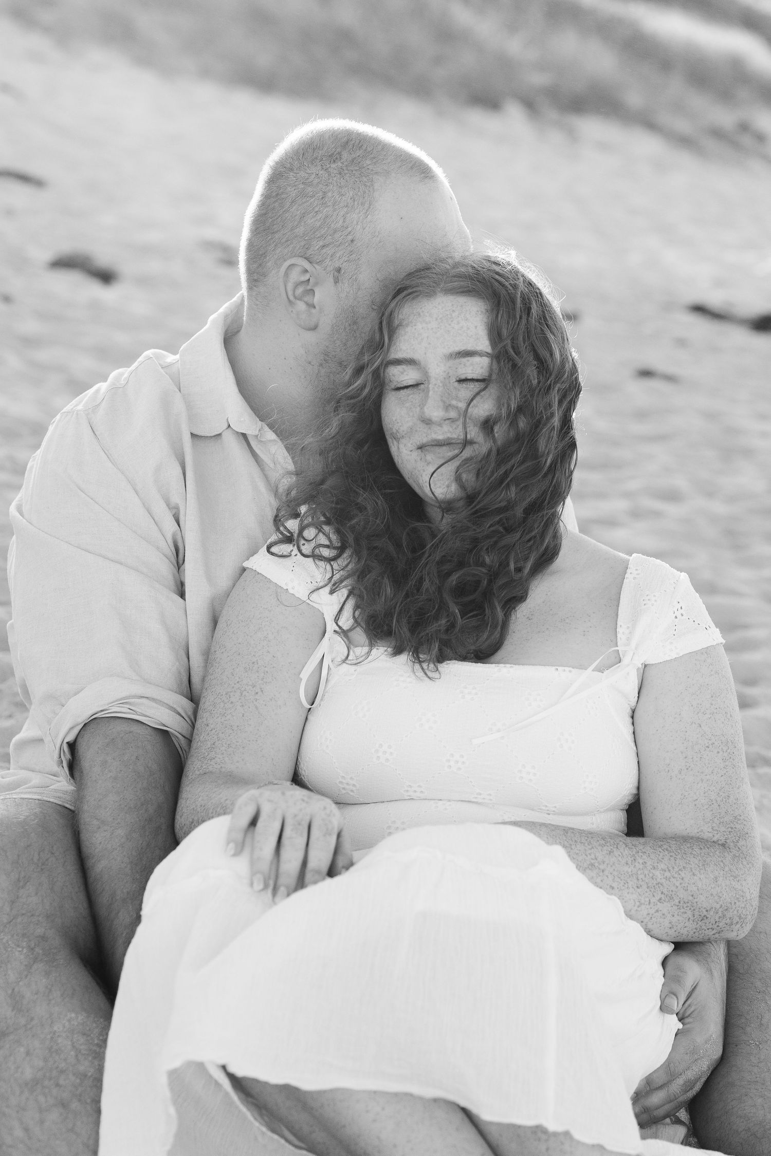 Black and white photo of a couple sitting on a beach, man kissing woman's head as she smiles with eyes closed.
