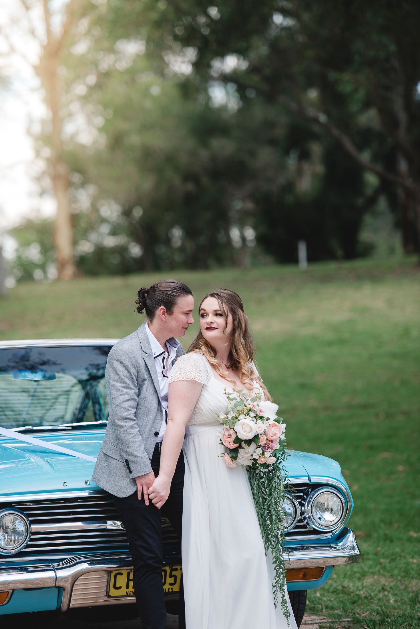 A newlywed couple poses with a vintage blue car while holding a floral bouquet.