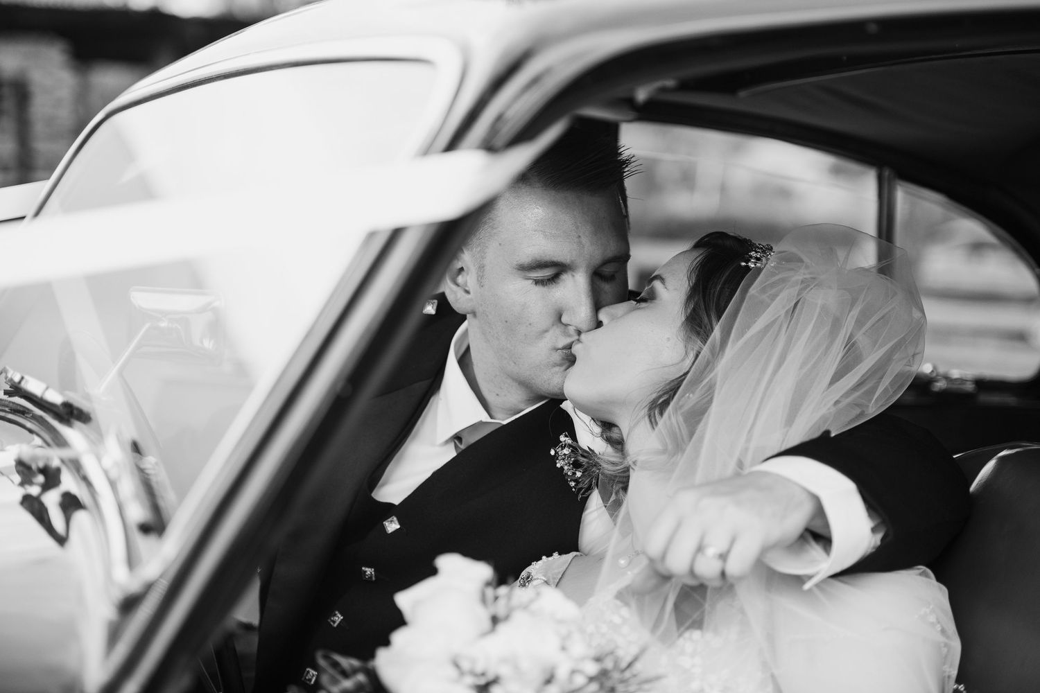 Black and white photo of newlyweds kissing in classic car after ceremony.
