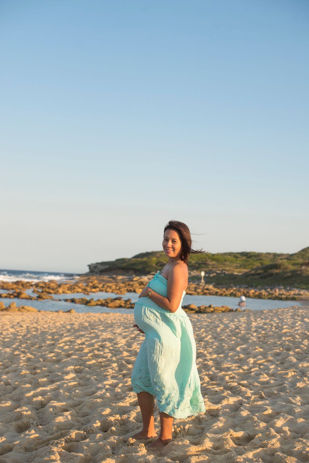 Mother to be in flowing mint dress poses on sandy beach during sunset photoshoot.