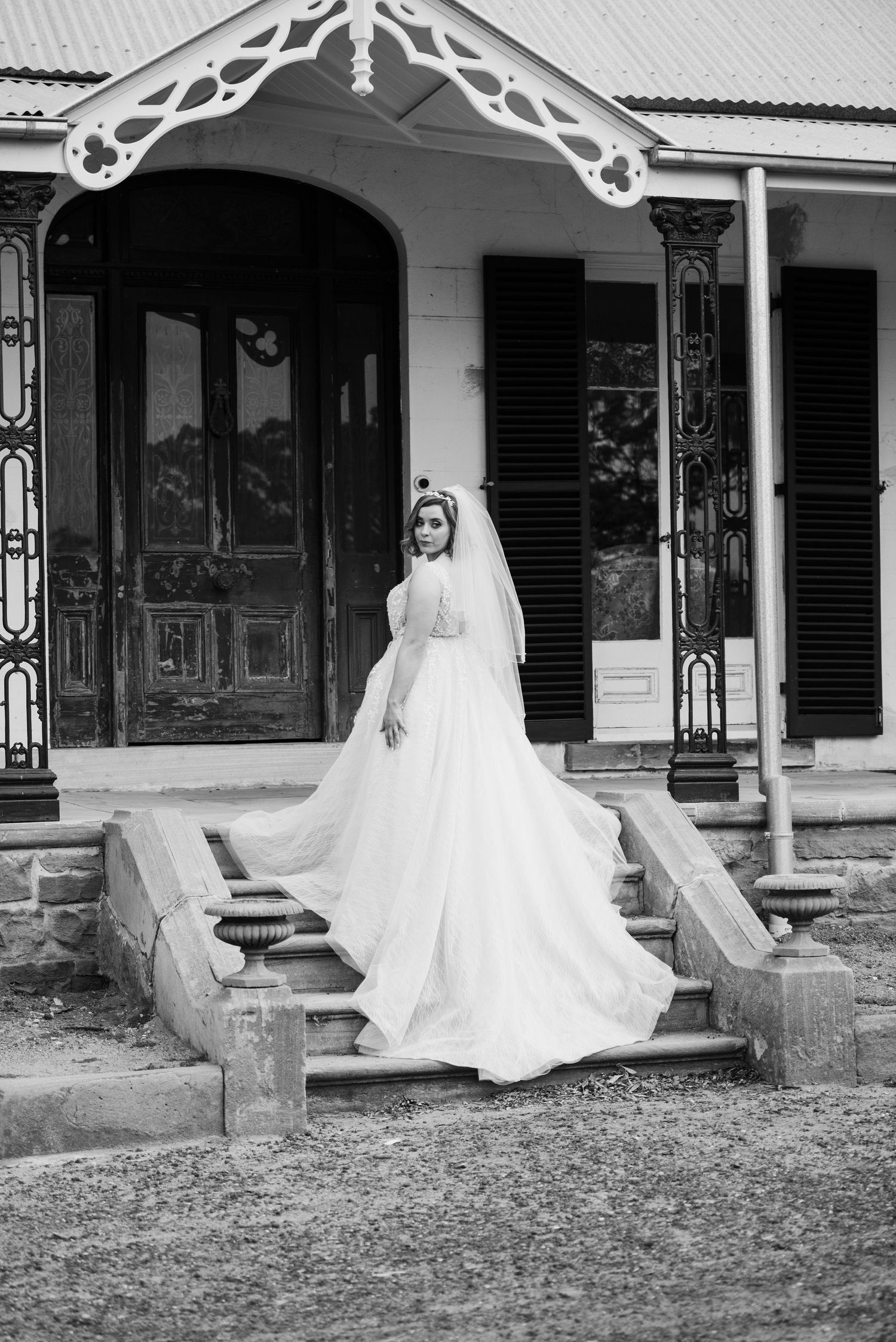 Black and white photo of a bride in a flowing white gown and veil standing on stone steps of a historic building.