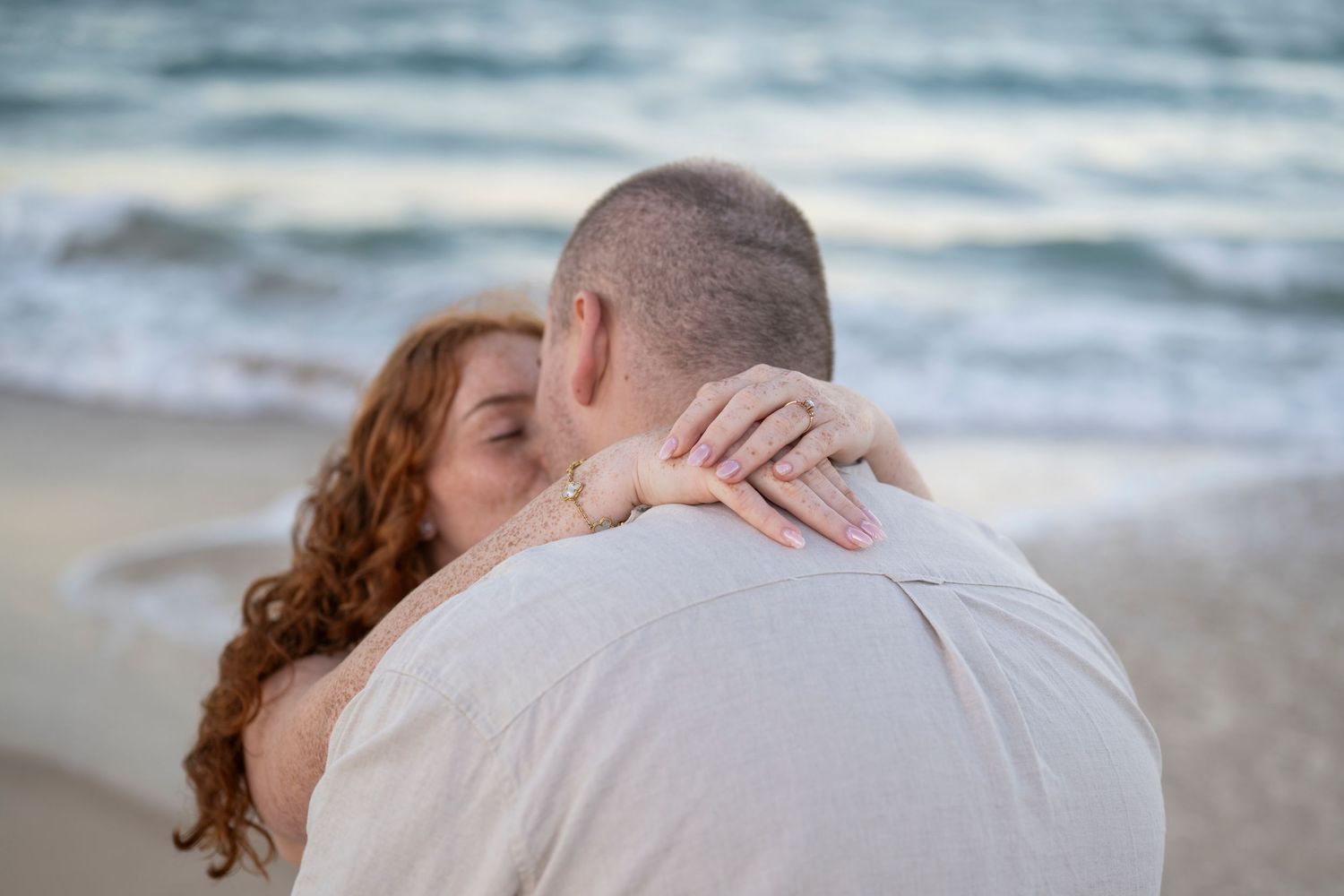 Couple embracing on a beach, woman with red curly hair showing engagement ring, ocean waves in background.
