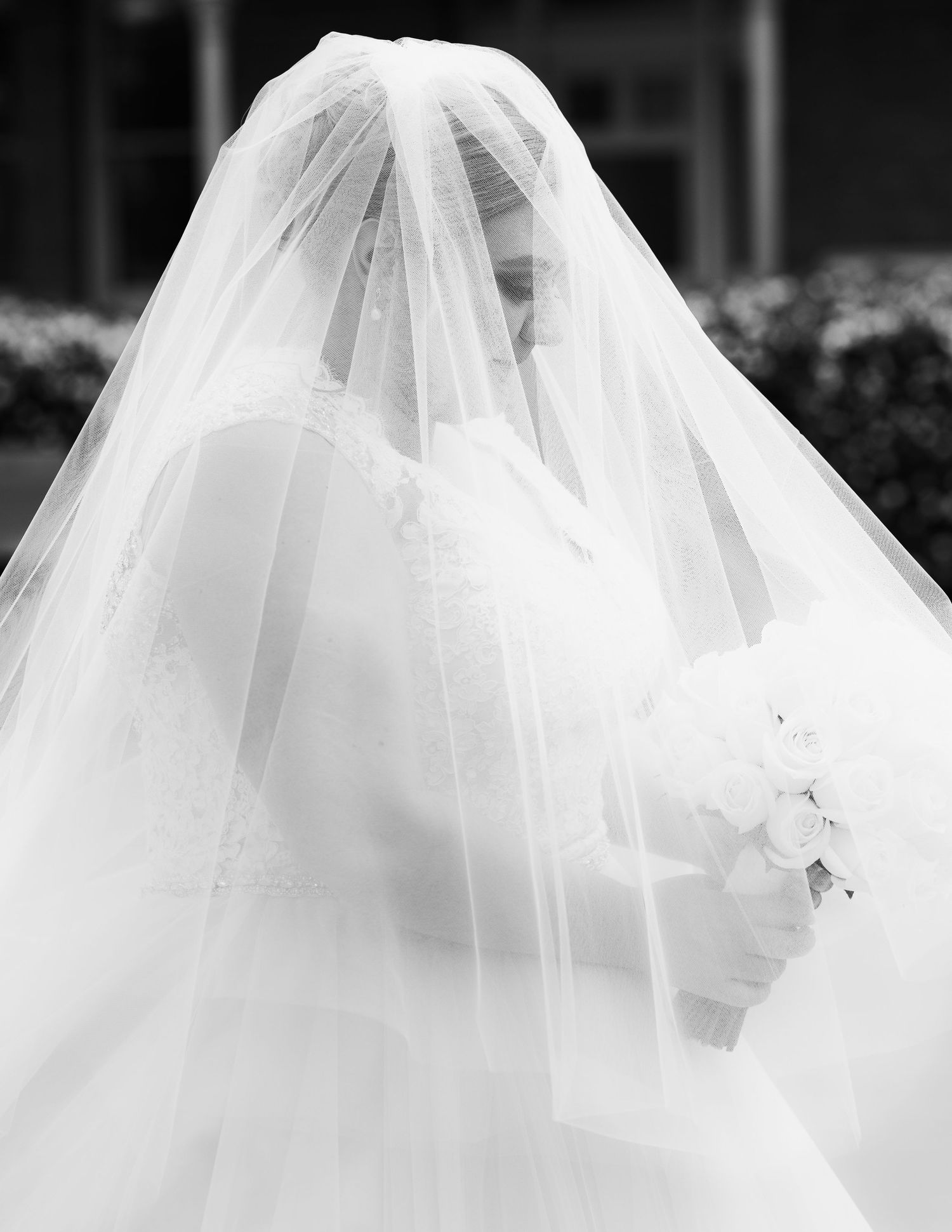 A bride wearing a flowing white veil stands elegantly in black and white photography.