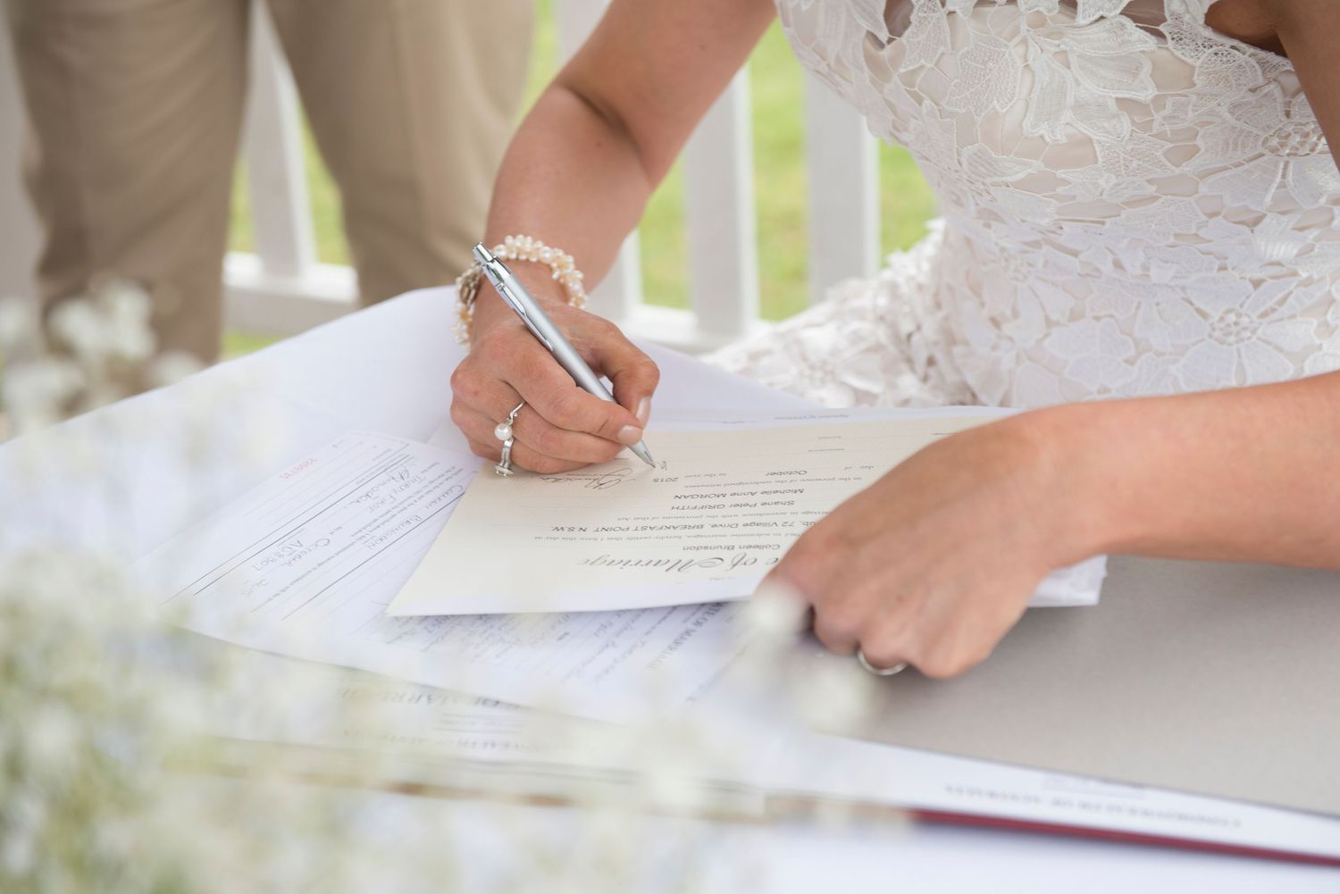 Close-up view of hands in white lace signing a document during a wedding ceremony.