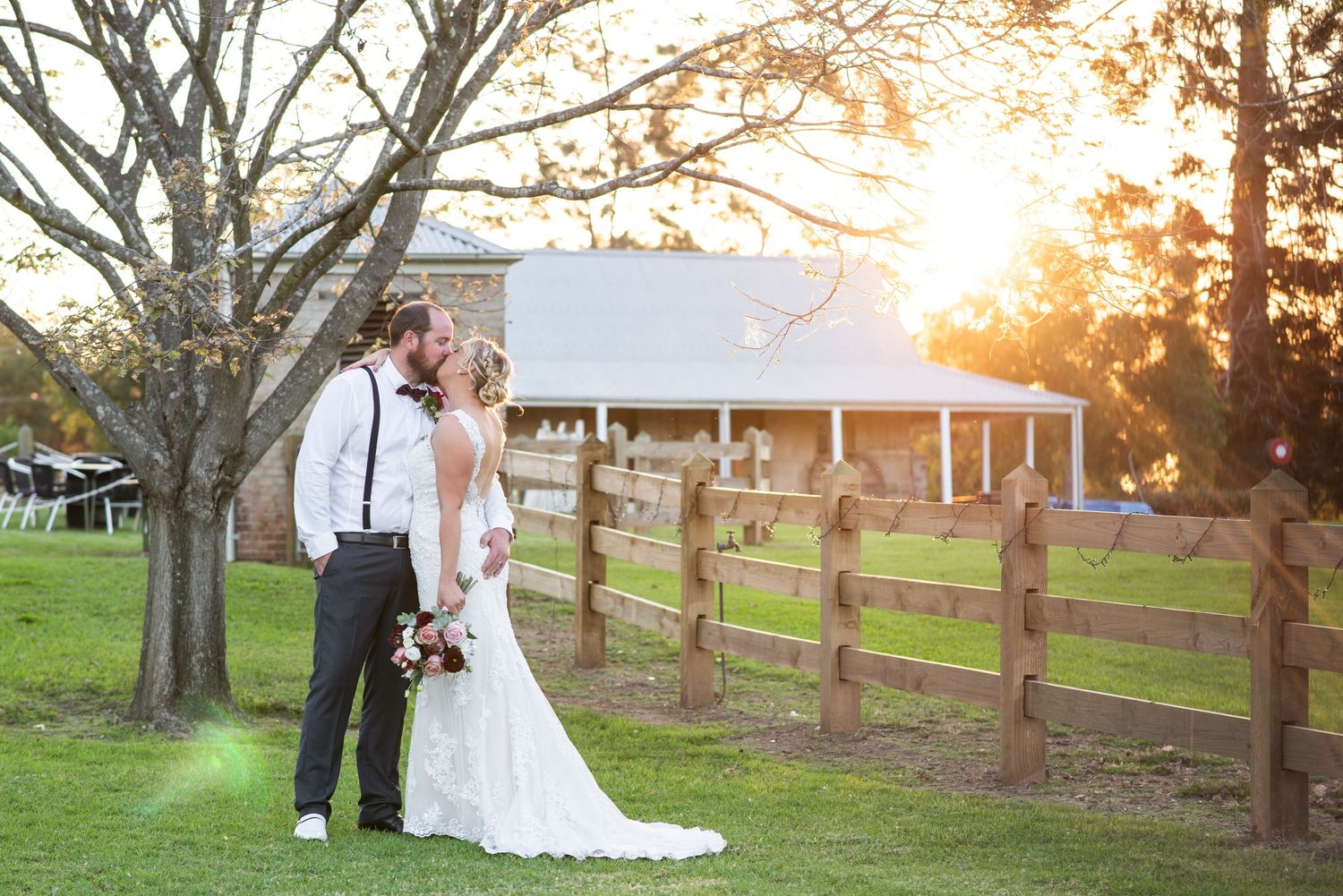 A couple poses for their wedding photos at sunset in front of a rustic barn and wooden fence.