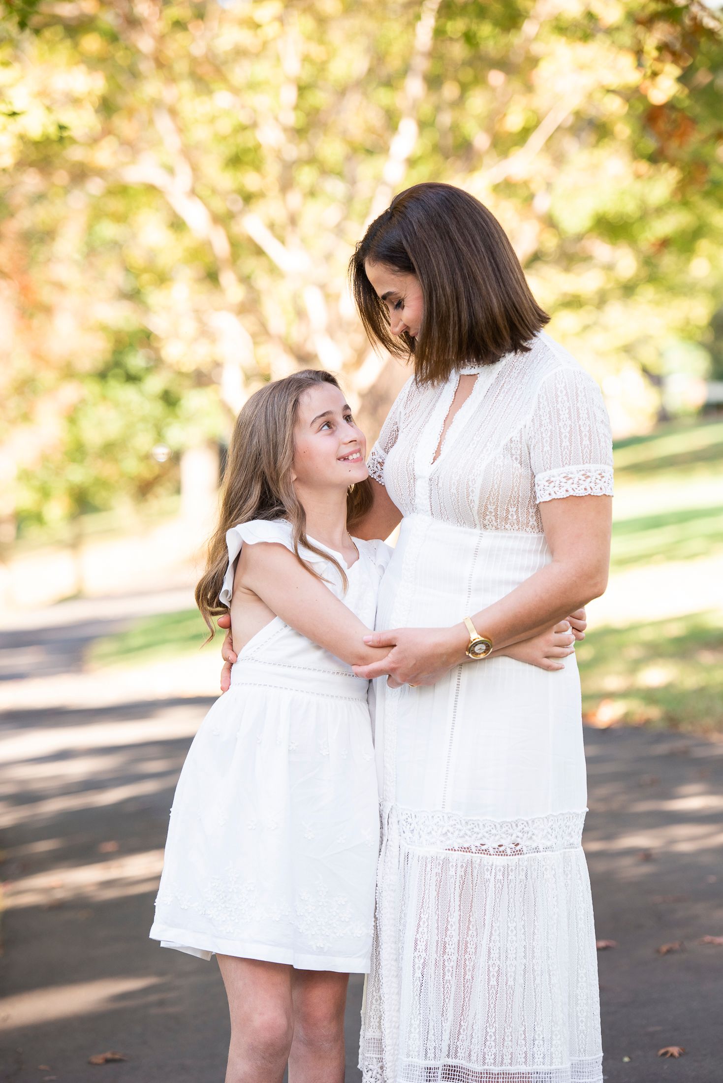 Mother and daughter in white dresses share intimate moments in park setting.