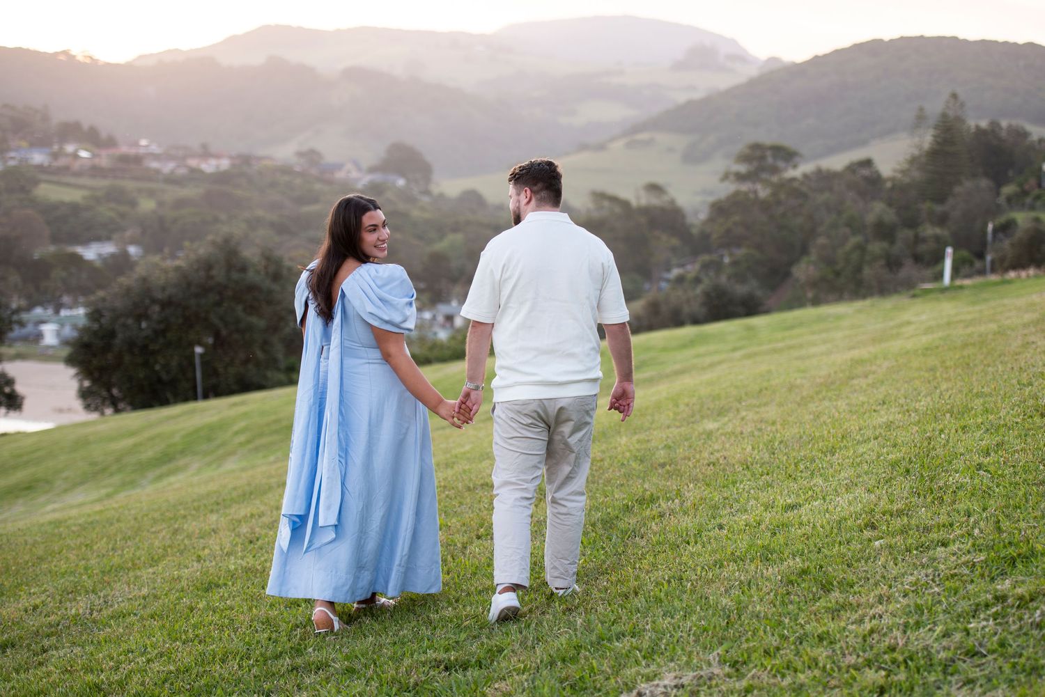 Couple in light blue and white outfits holding hands while walking on a grassy hillside in Kiama at sunset.