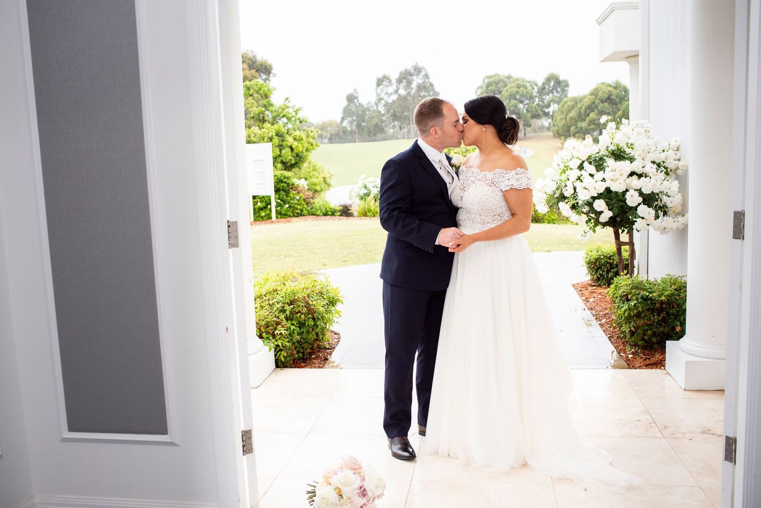 A couple shares a romantic moment during their wedding on a covered porch with white columns and greenery.