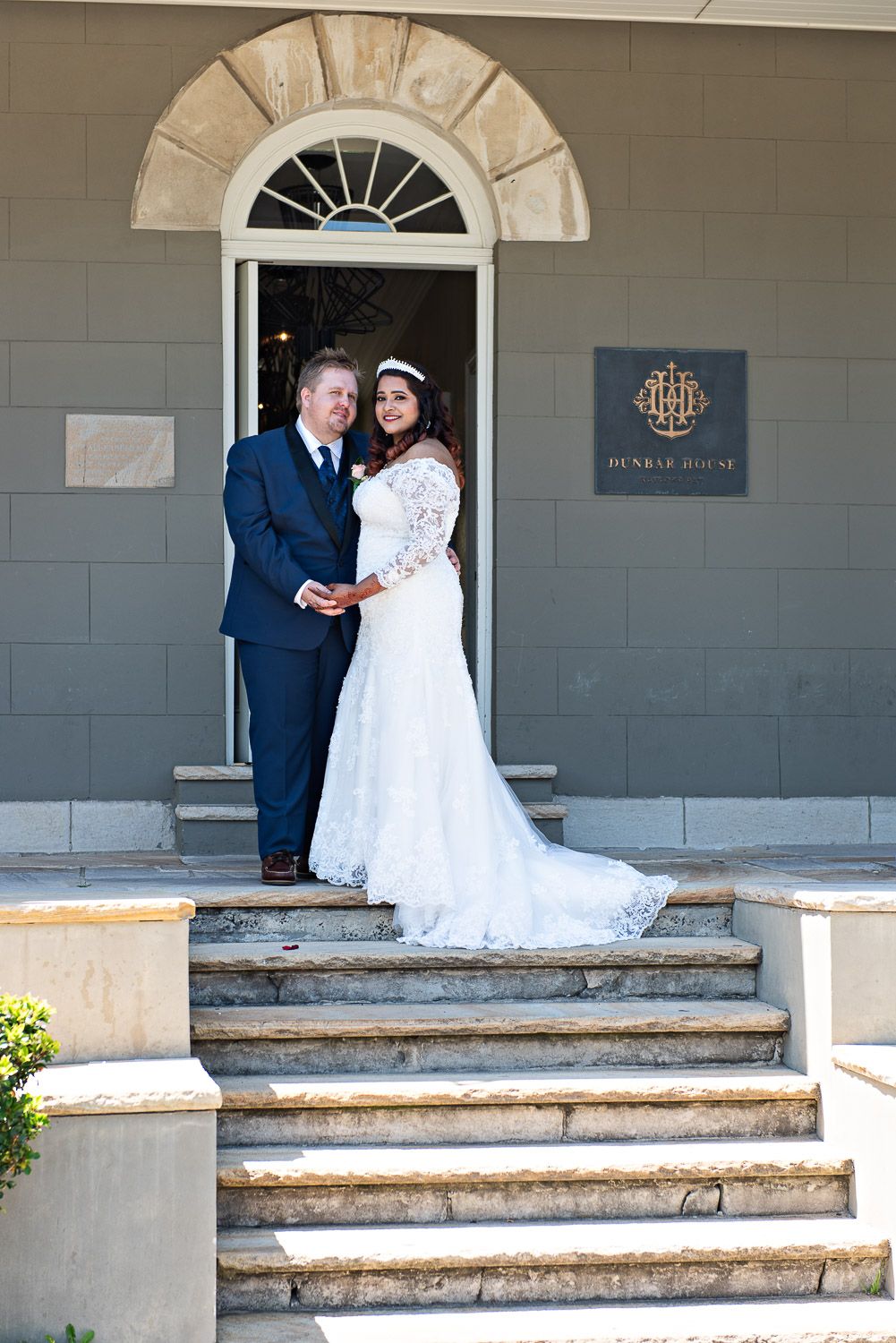 A couple in wedding attire shares an intimate moment on stone steps outside Dunbar House with arched doorways.