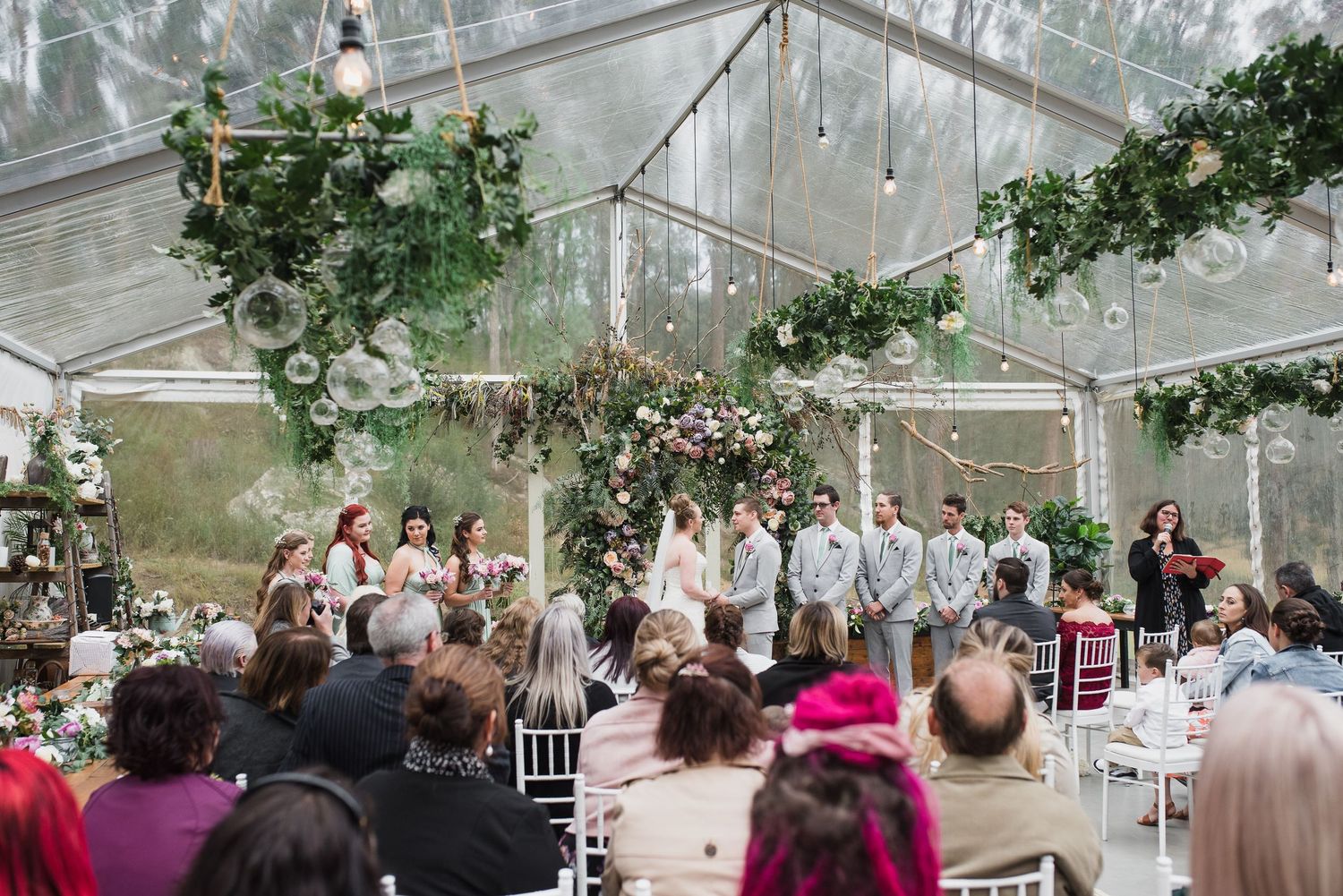 A wedding ceremony takes place inside a clear room with white chairs arranged in rows and greenery hanging overhead.
