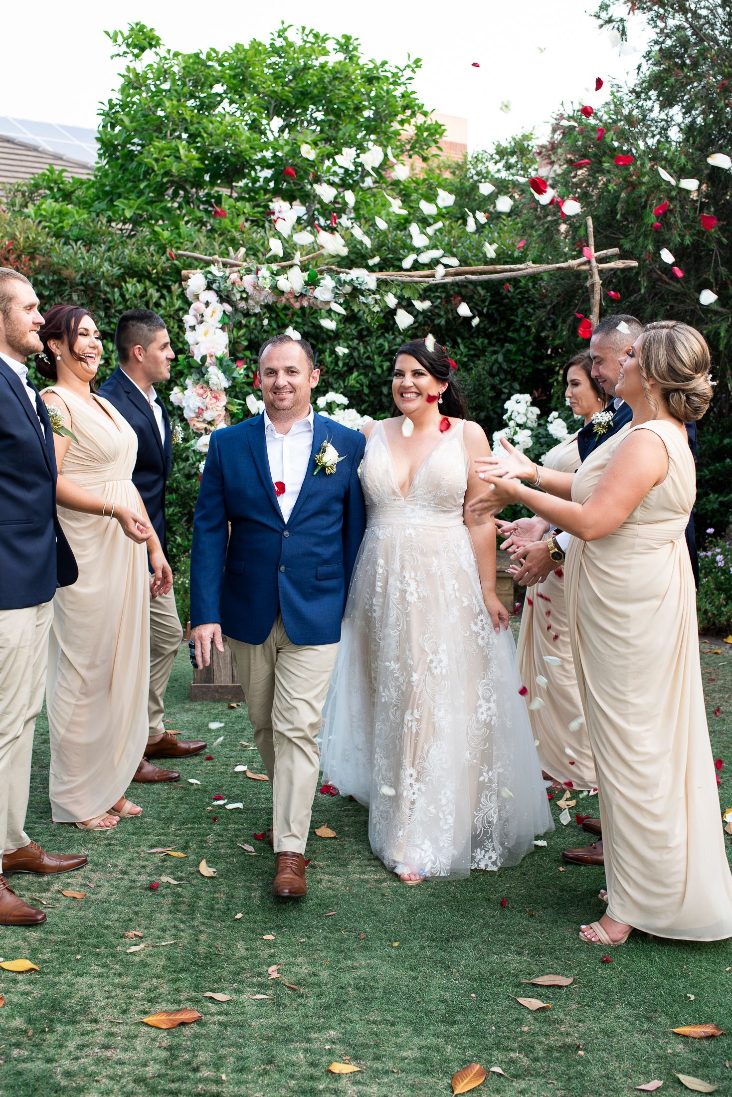 Wedding party in navy and beige attire poses together on brick pathway surrounded by lush greenery.