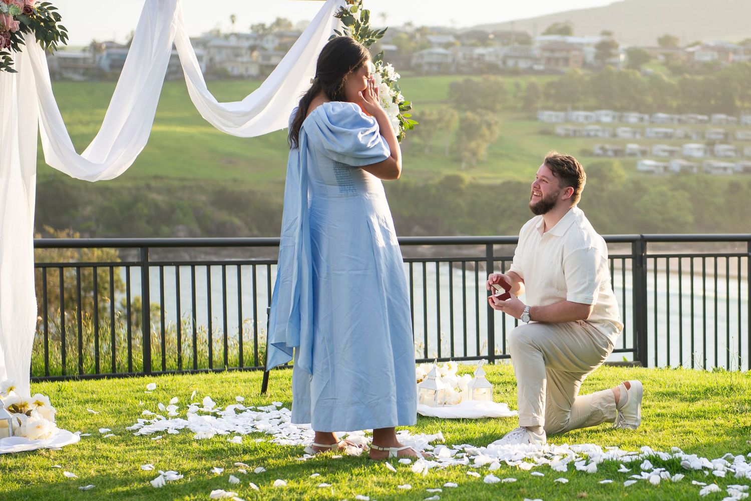 A couple shares a tender moment during a surprise proposal in Kiama with white decorations and scenic views.