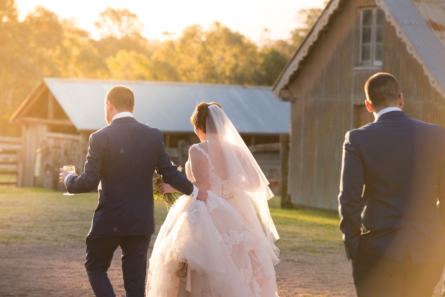 Wedding couple strolls together during golden sunset with rustic buildings in background.