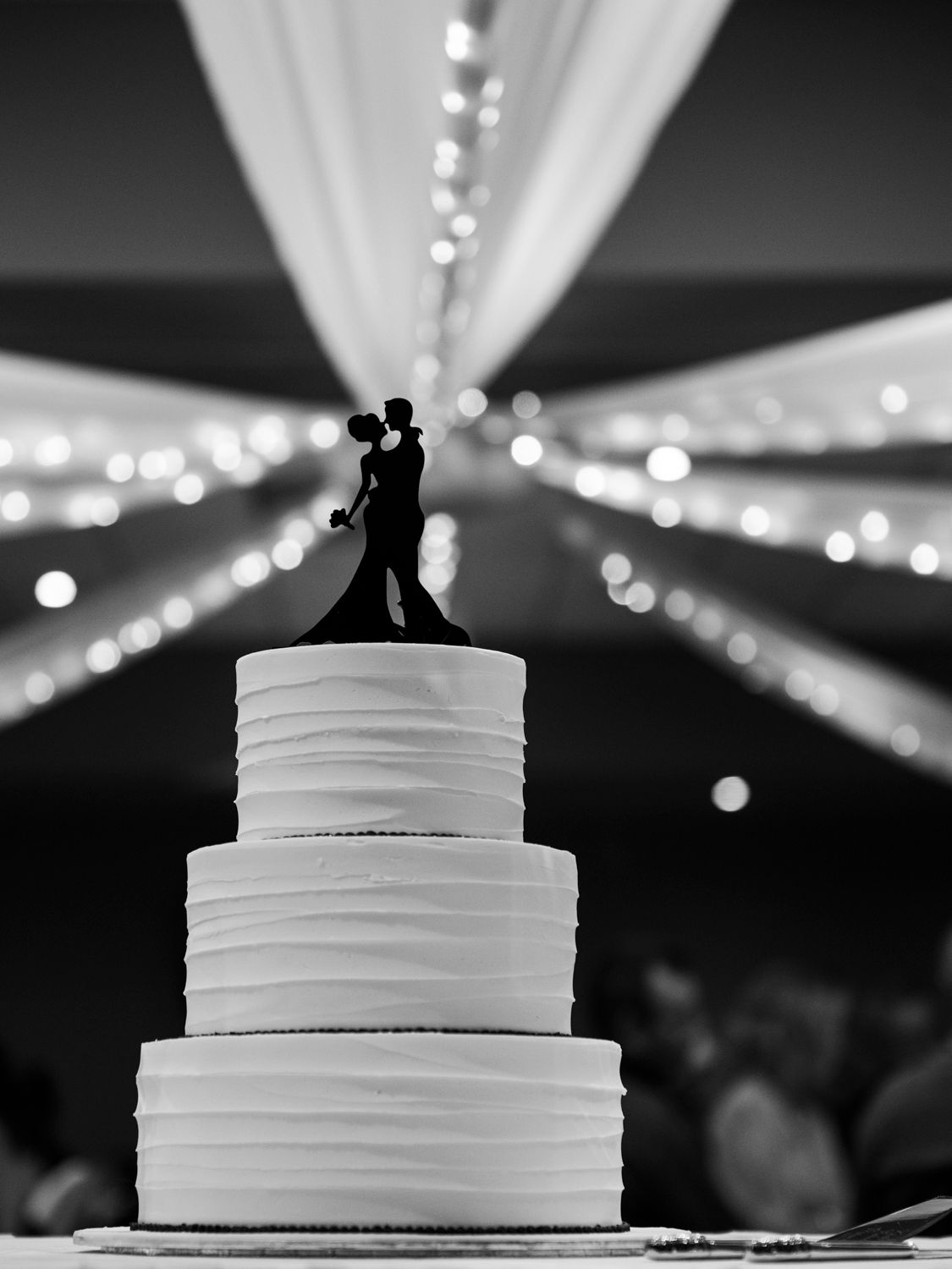 Elegant white wedding cake with tiered layers under twinkling reception lights.