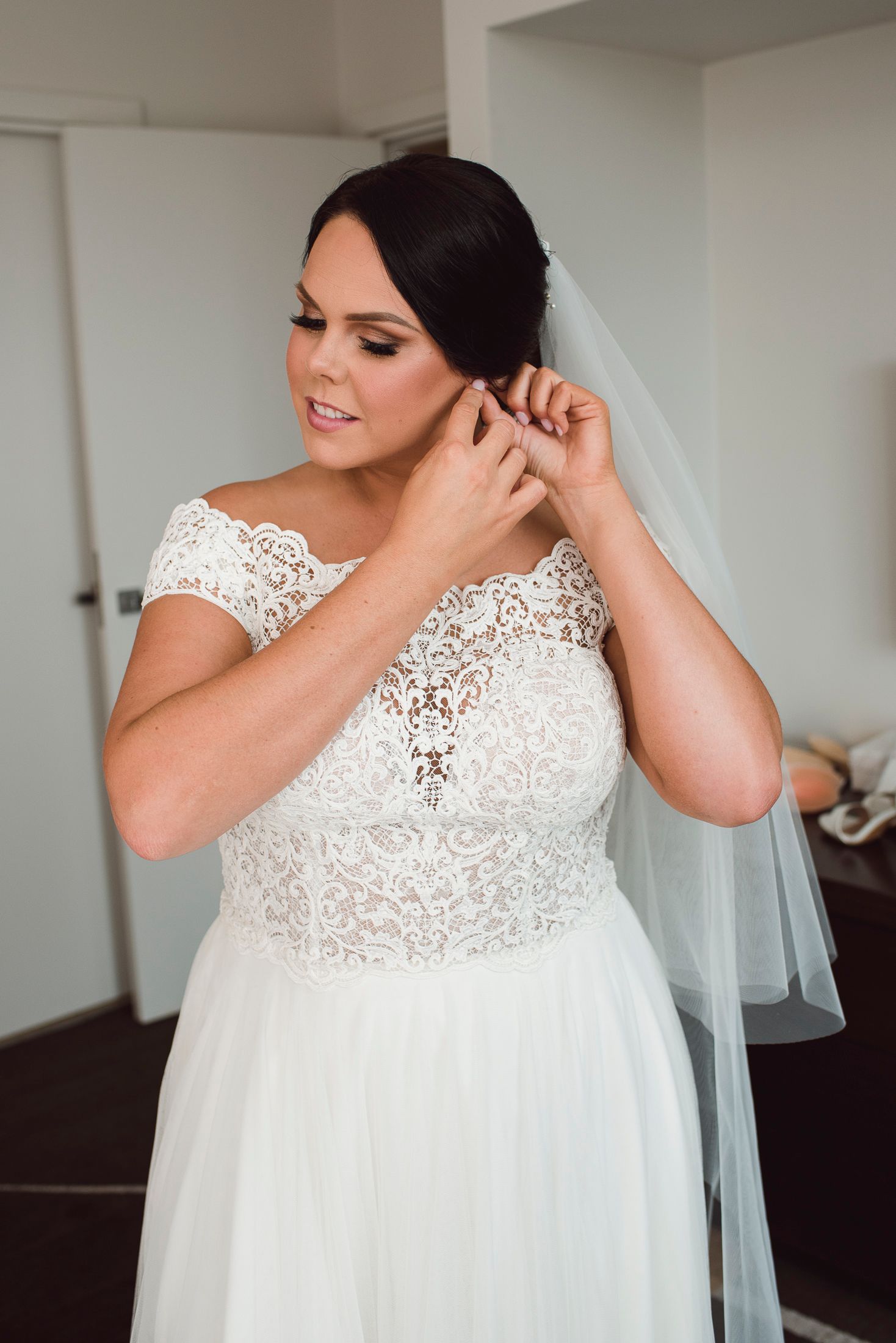A bride in an elegant lace-bodice wedding gown with cap sleeves puts on earrings while wearing a flowing veil.