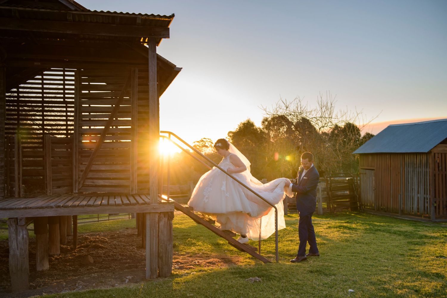 A couple in wedding attire shares a romantic moment at sunset near a rustic wooden barn structure.