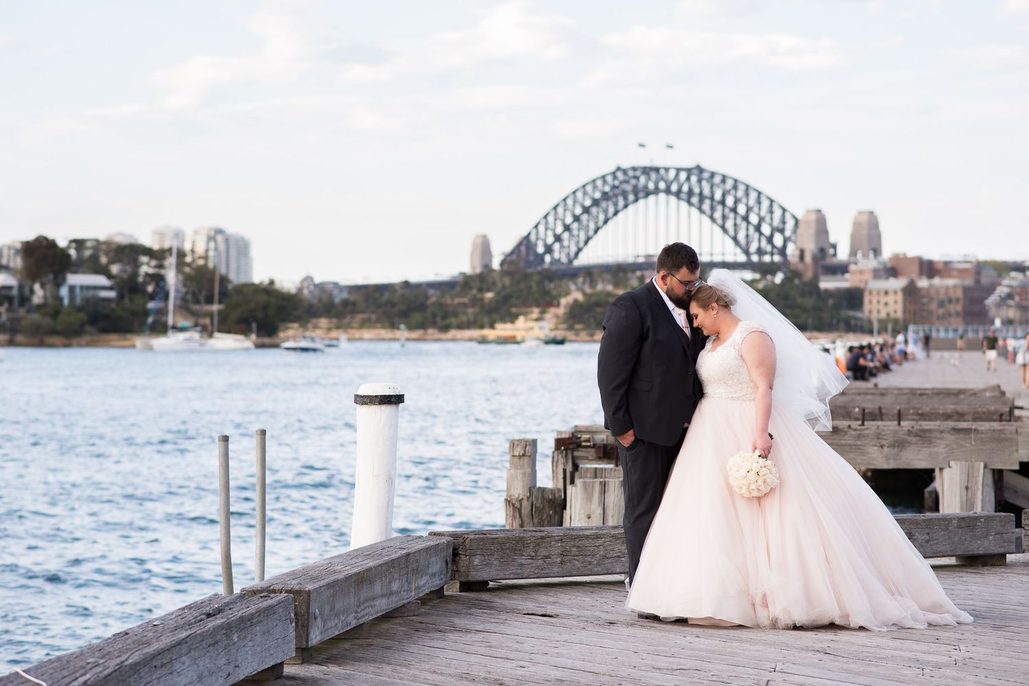 A couple shares a romantic moment on a pier with the iconic Sydney Harbour Bridge in the background.
