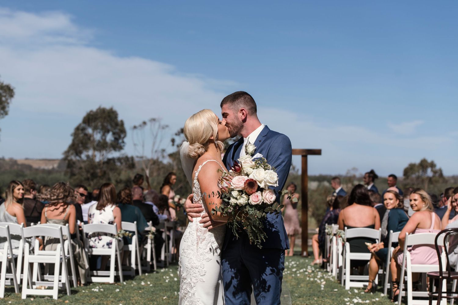 Wedding ceremony in progress with guests seated in white chairs on a sunny outdoor lawn.