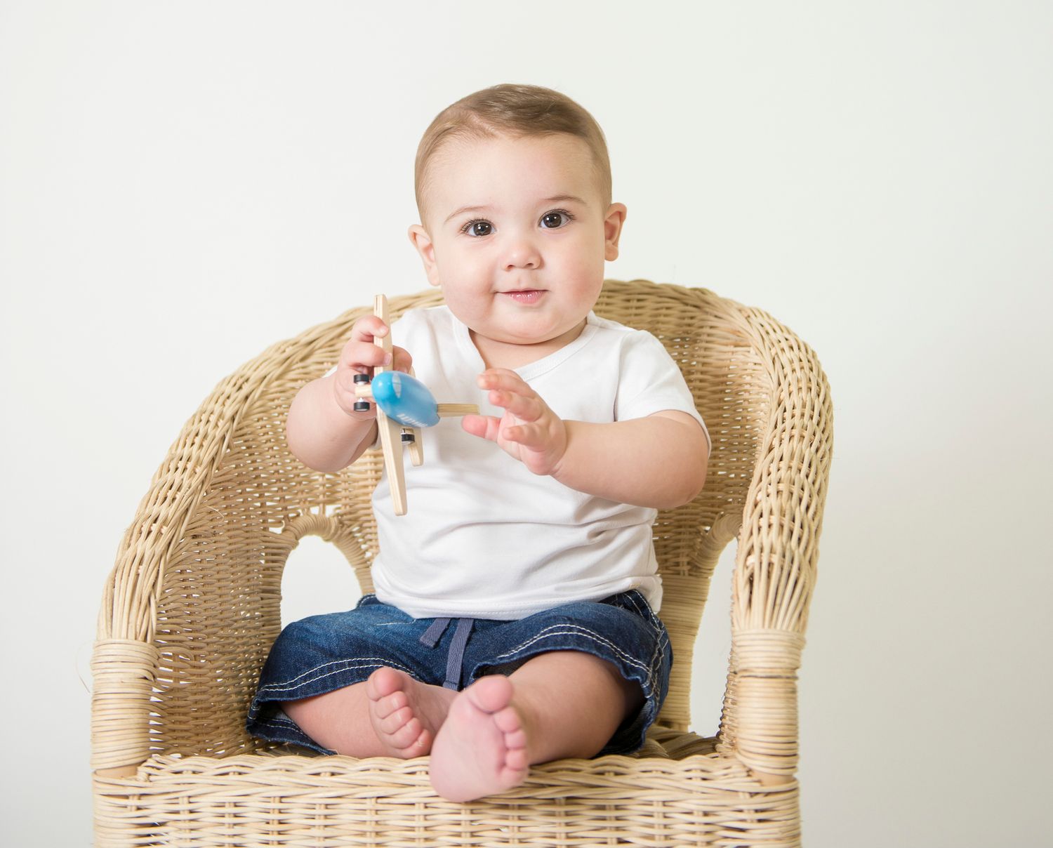 Baby sitting in wicker chair wearing white onesie and blue pants.