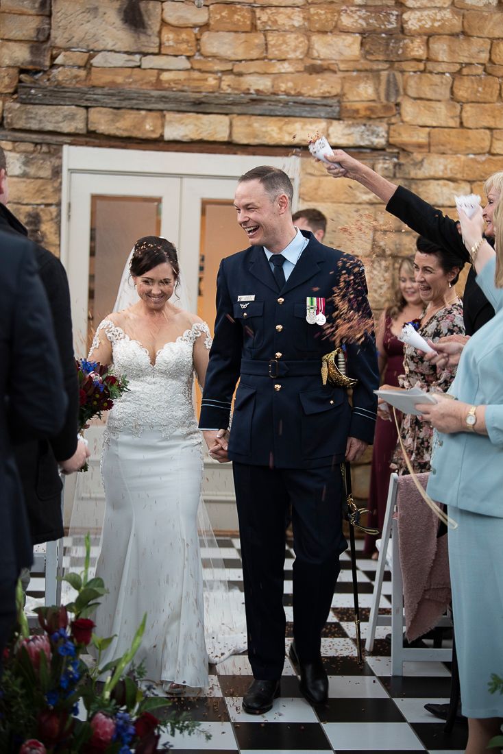 A wedding couple walks down an aisle while guests throw flower petals in celebration at a stone venue.