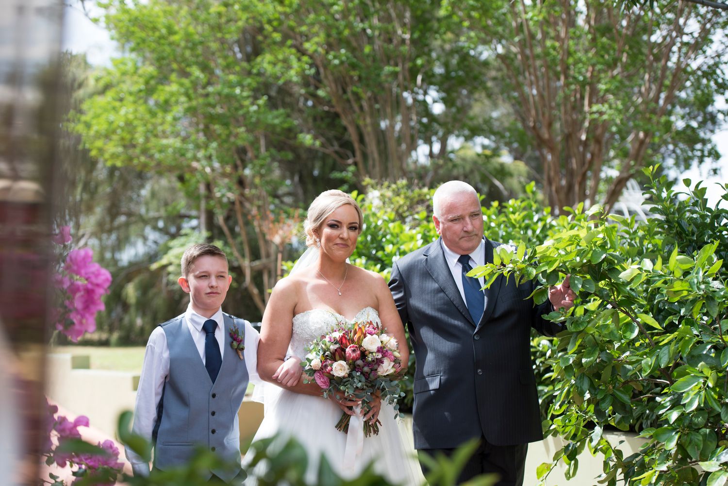 Bride with son and father on her way to the wedding ceremony in a lush garden setting.