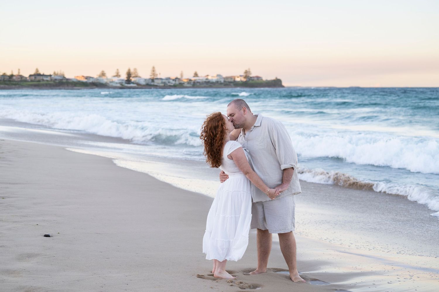 Couple kissing on a sandy beach at sunset, wearing white and grey outfits, with ocean waves in the background.