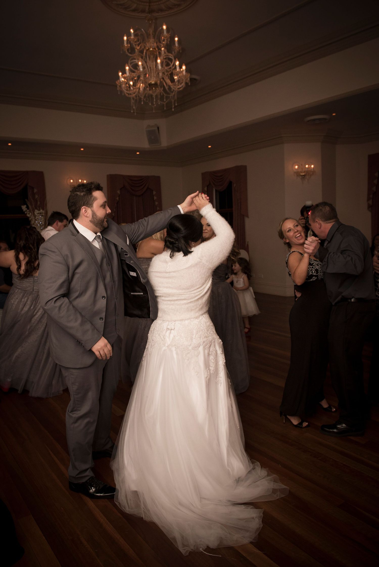Dancing together on a dark wooden dance floor beneath elegant chandeliers.